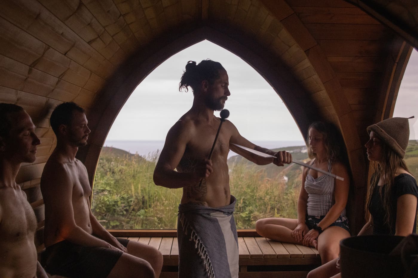 Guide leading a Soul Sweats soundbath inside the arched-window sauna, using a mallet and singing bowl while guests relax on the benches.