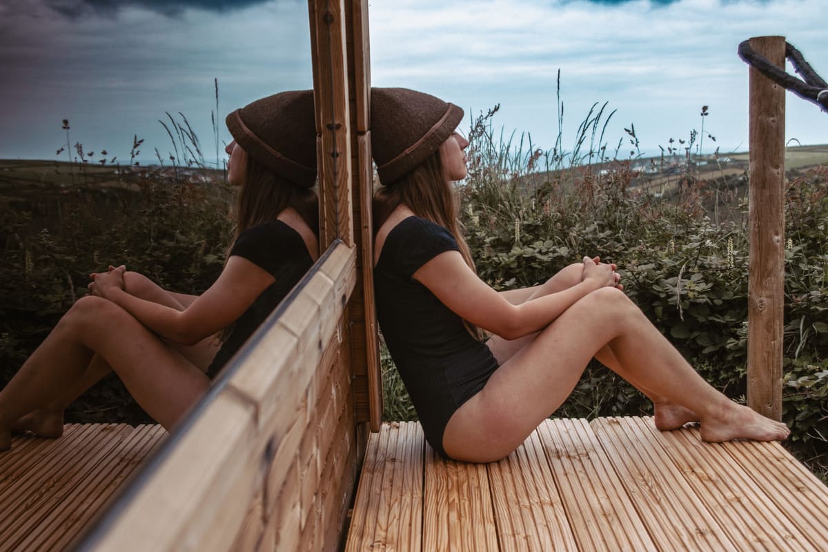 Woman sitting in quiet reflection outside the Soul Sweats sauna, leaning against the mirrored timber wall with coastal valley views in the background.