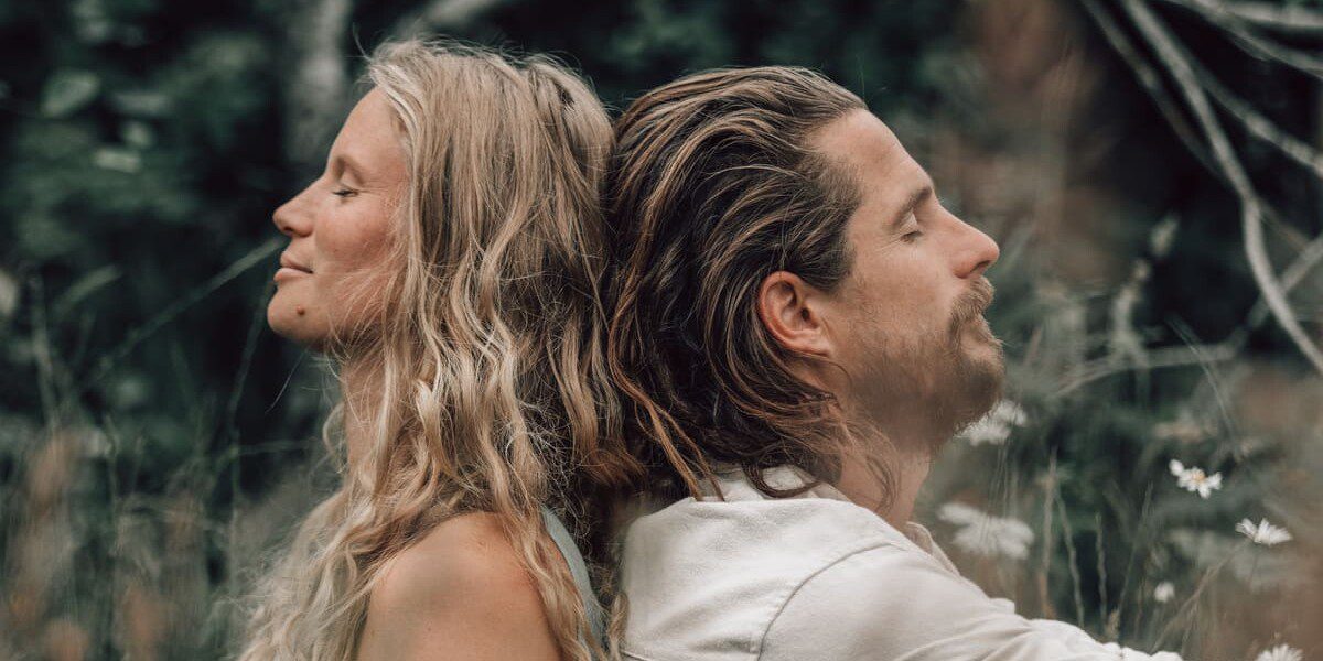Jana and Sam, co-owners of Soul Sweats, sitting back-to-back outdoors in nature, smiling softly with wild greenery behind them.