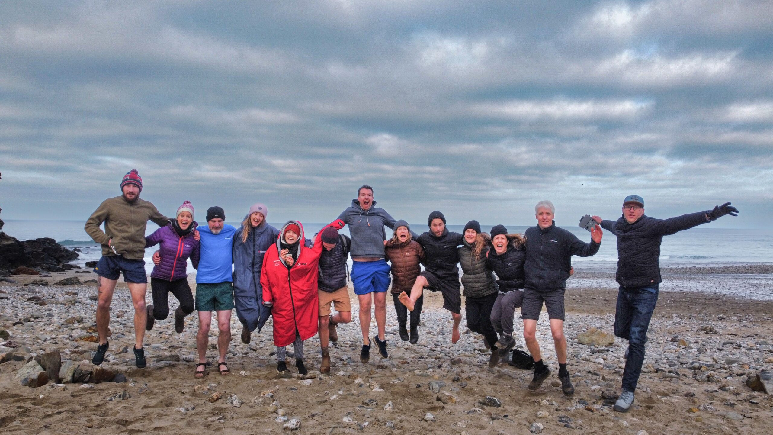 Soul Sweats retreat group jumping together on Porthtowan Beach, smiling in the wind with waves and dramatic clouds behind them.
