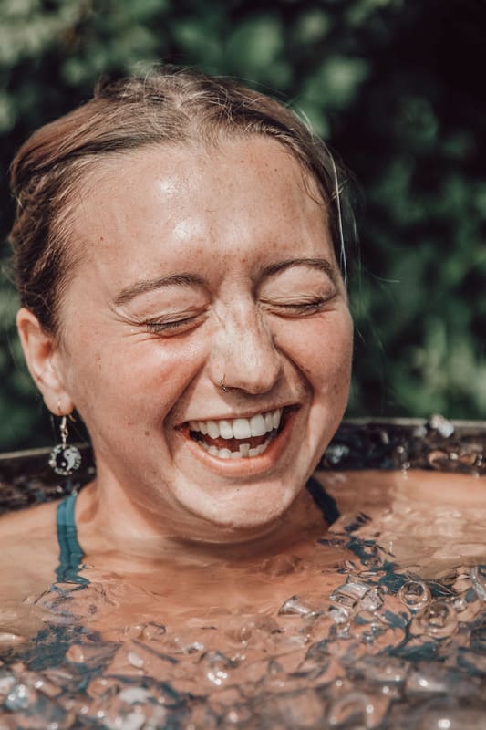 Woman smiling with her eyes closed during a Soul Sweats cold-water immersion, surrounded by ice and greenery in the background.
