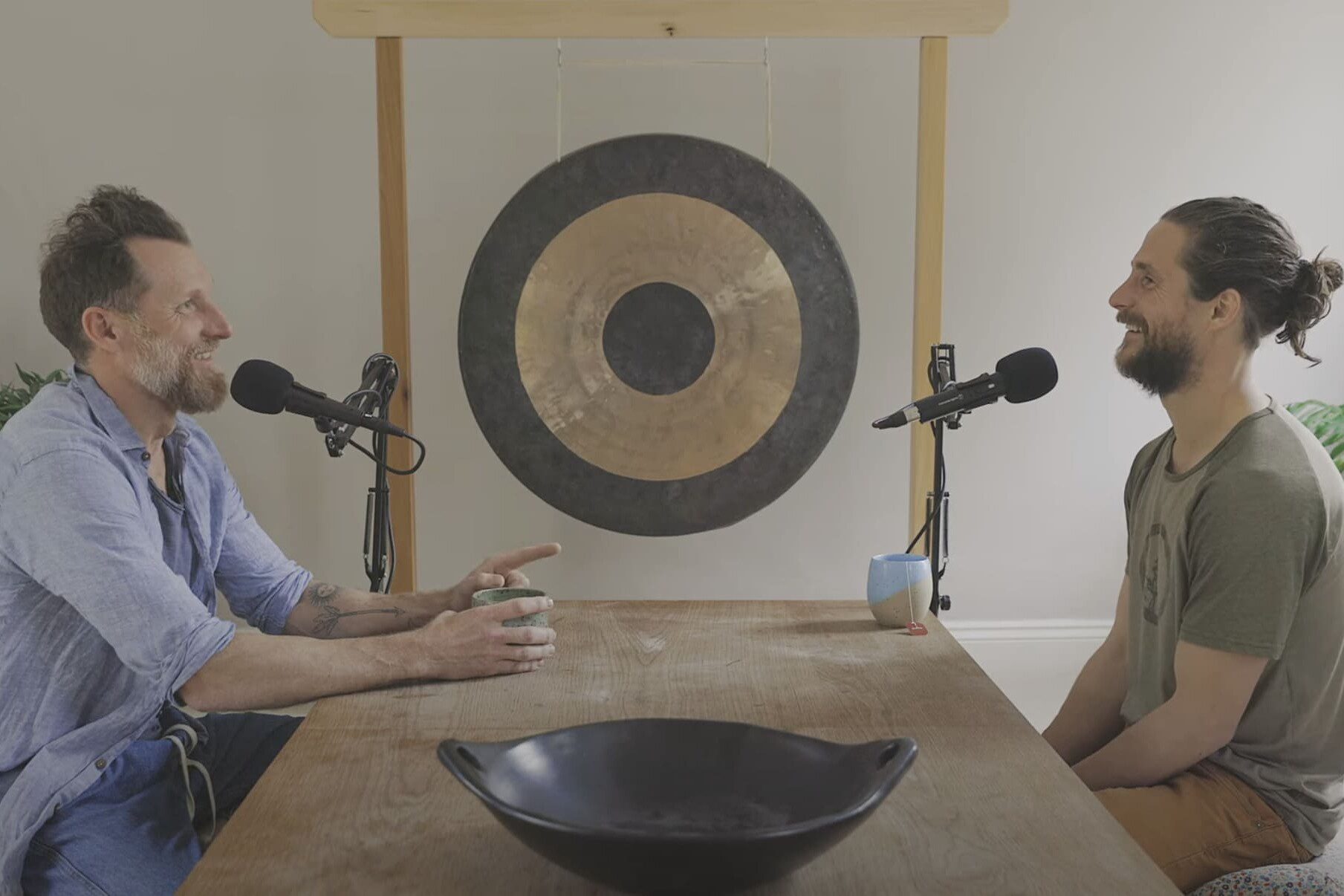 Two men recording a podcast conversation at a wooden table, seated in front of a large gong in a calm, minimalist studio space.