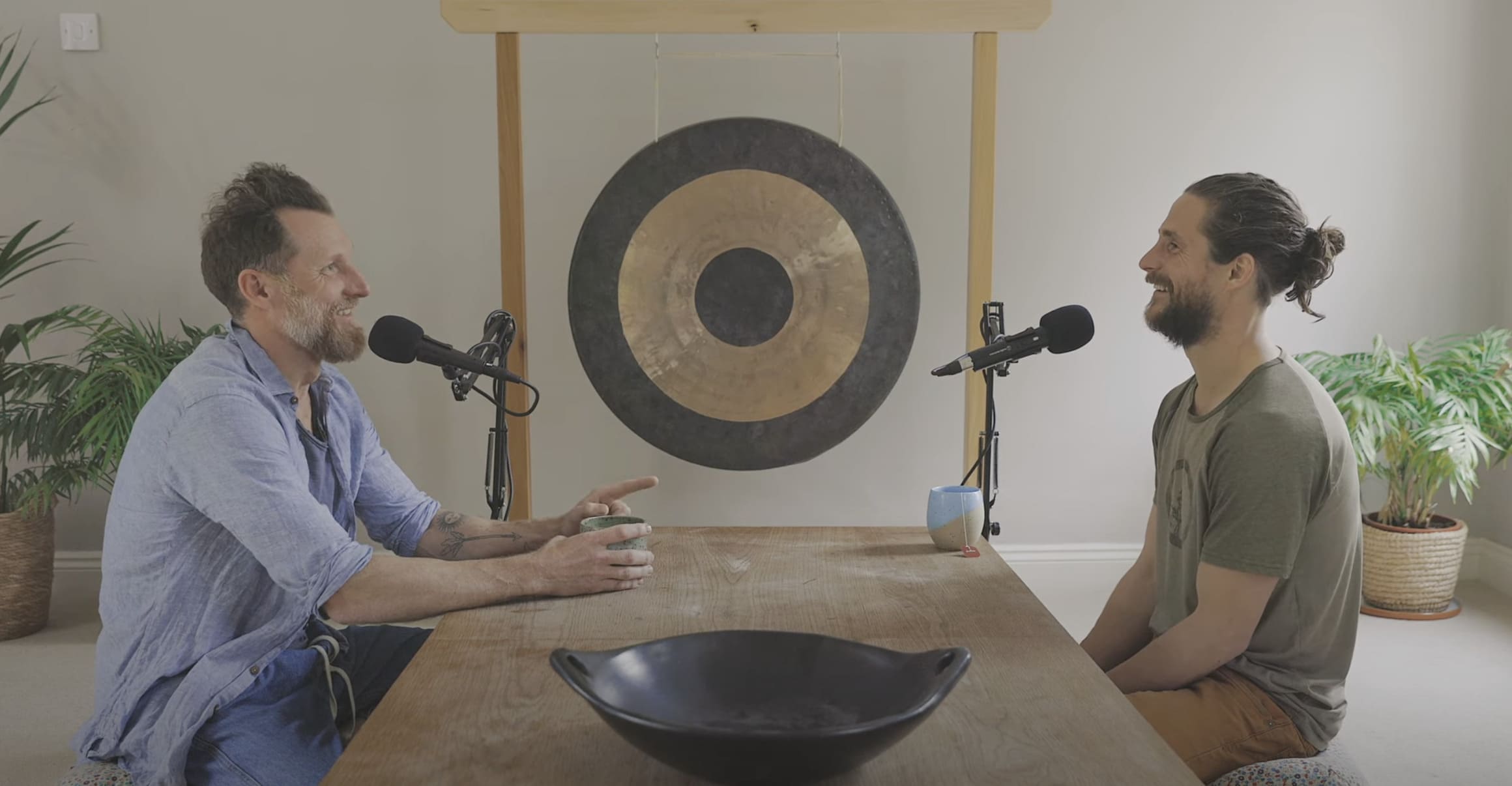 Two men recording a podcast conversation at a wooden table, seated in front of a large gong in a calm, minimalist studio space.