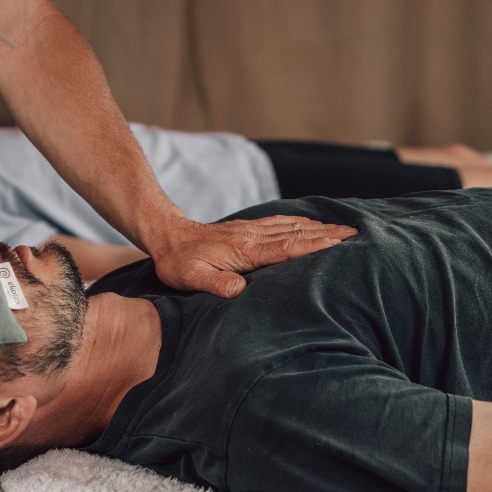 Practitioner gently placing their hand on a man’s chest during a Soul Sweats holistic healing session, while he rests with an herbal eye pillow.