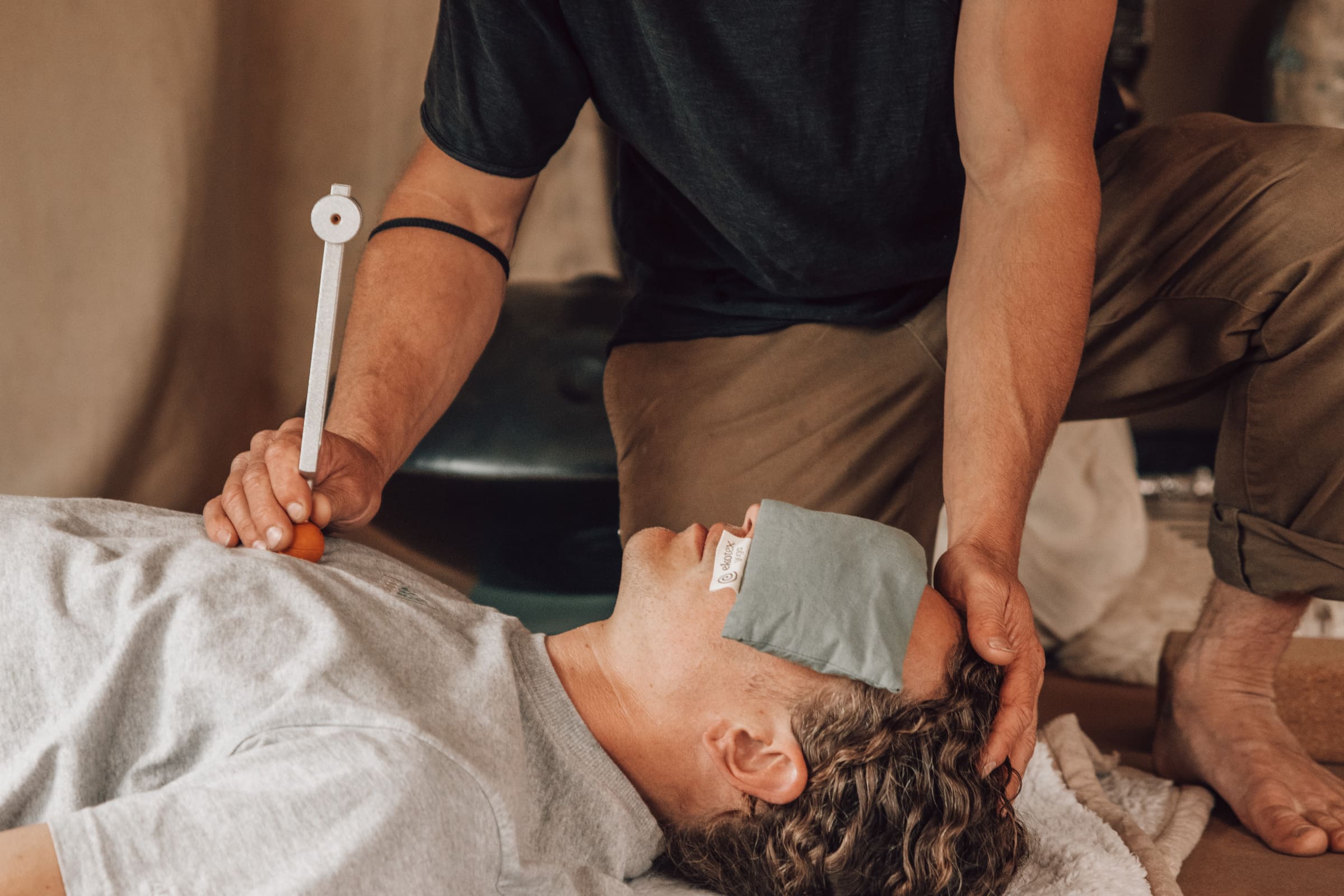 Practitioner holding a tuning fork and gently supporting a client’s head during a Soul Sweats holistic healing session, with the client resting under an eye pillow.