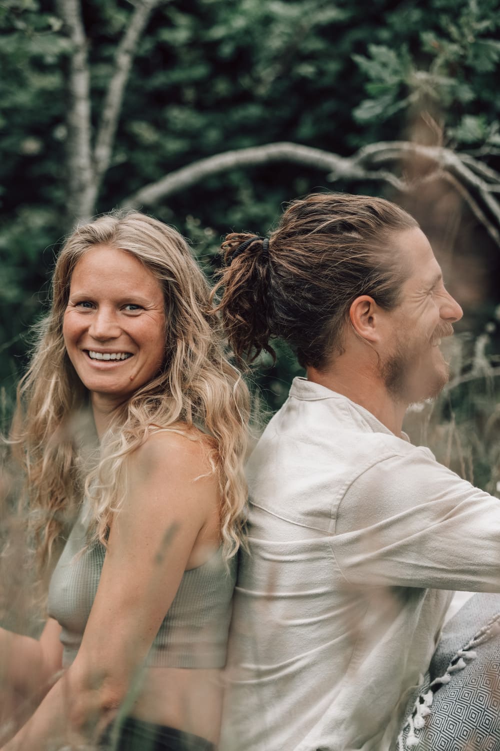 Sam and Jana, co-owners of Soul Sweats, sitting back-to-back outdoors and smiling in the middle of lush green woodland.