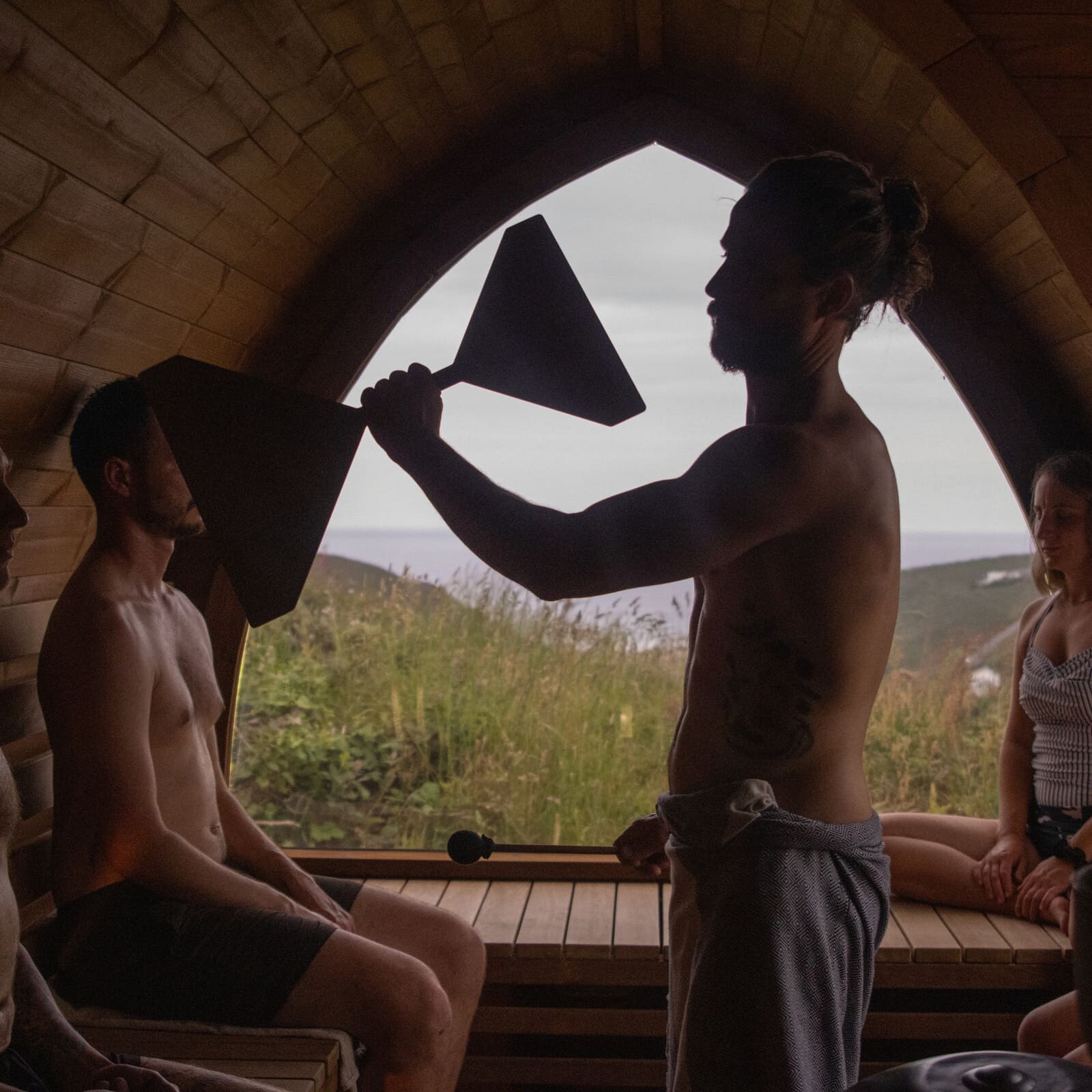 Guide leading an Aufguss ritual inside the Soul Sweats sauna, wafting steam with towels as guests sit around the arched window overlooking the Cornish landscape.