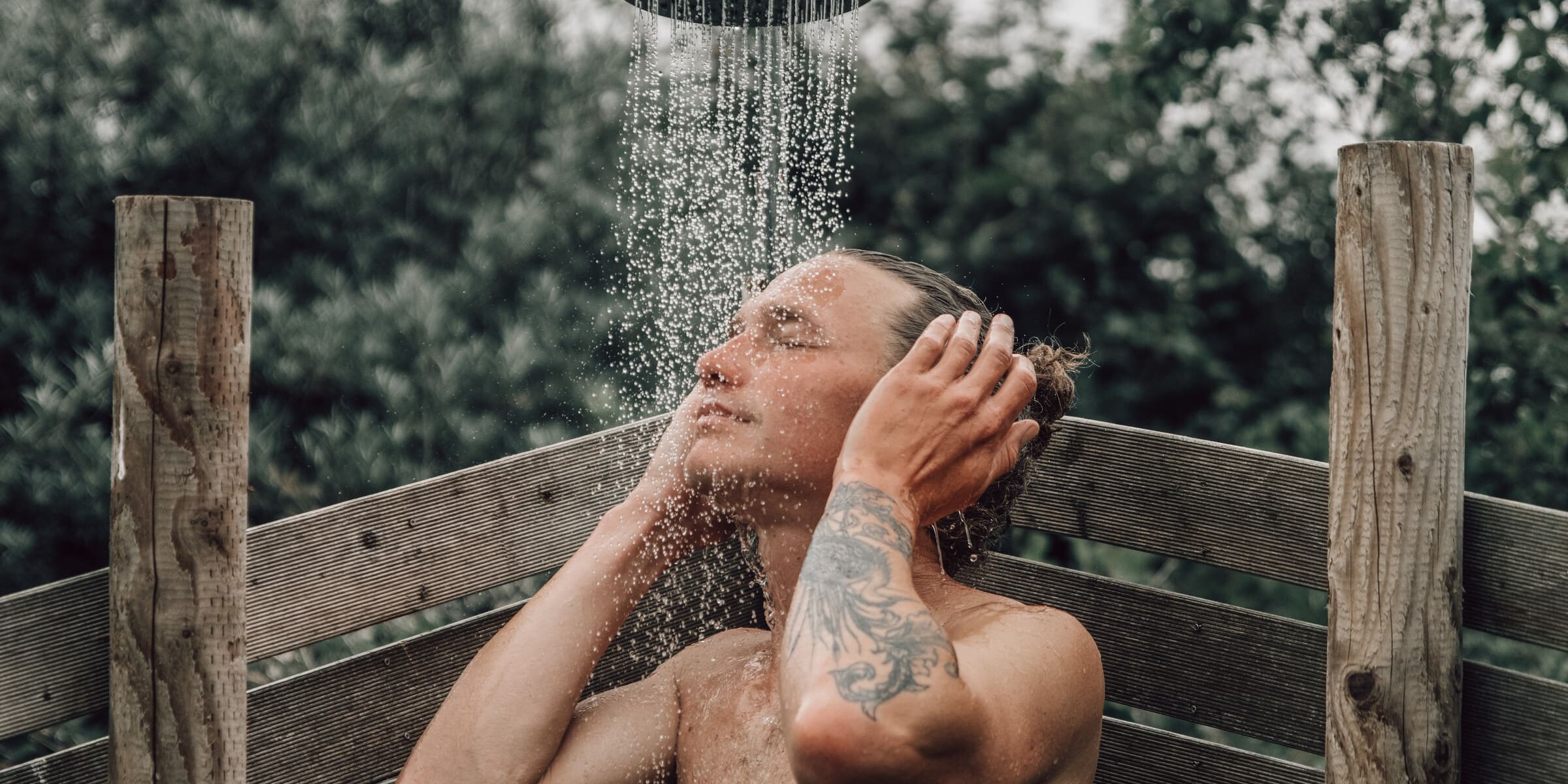 Man standing under an outdoor cold shower at Soul Sweats, eyes closed with hands on his head as water cascades down in a wooden shower enclosure.