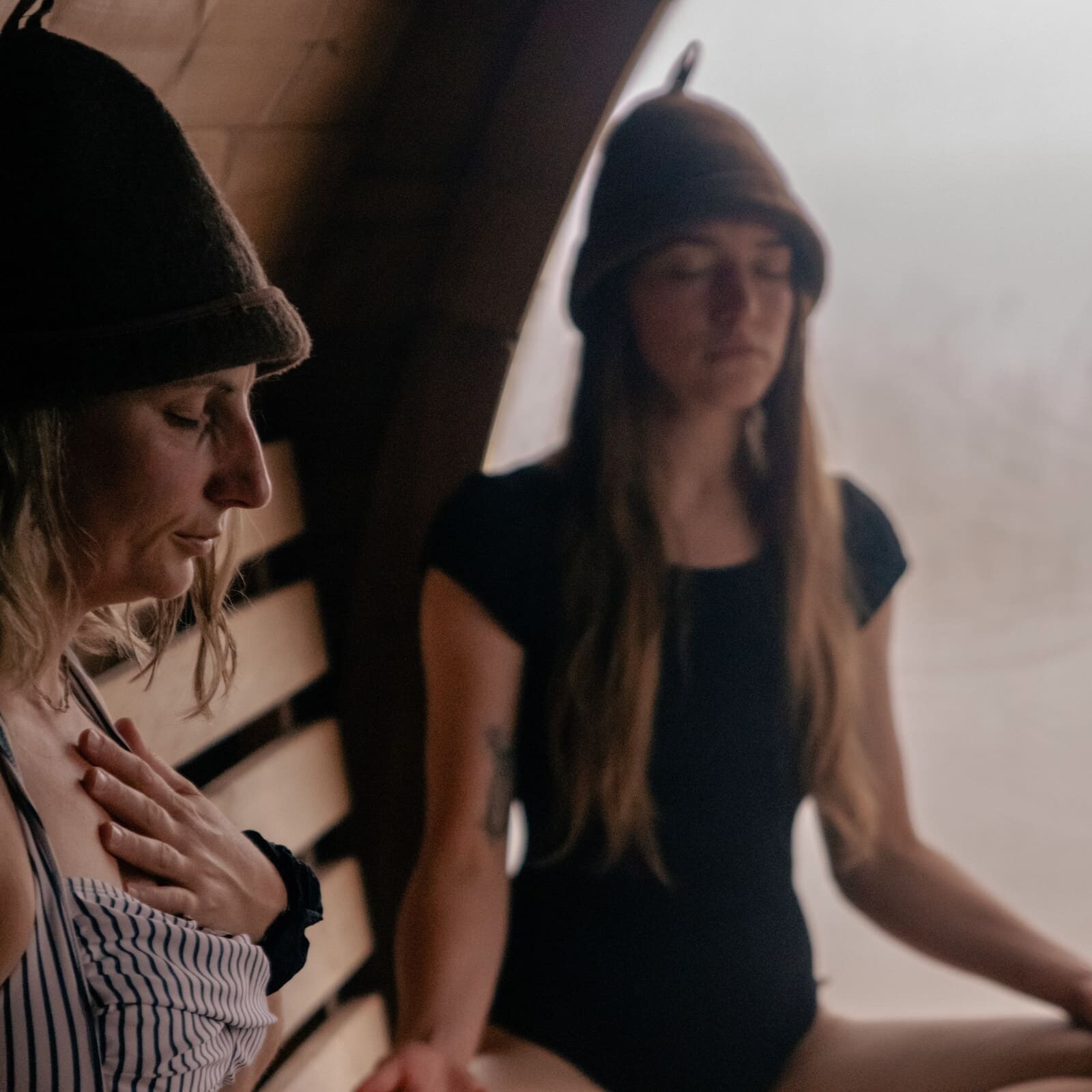 Two women meditating inside the Soul Sweats sauna, wearing felt sauna hats and sitting in calm, warm light.