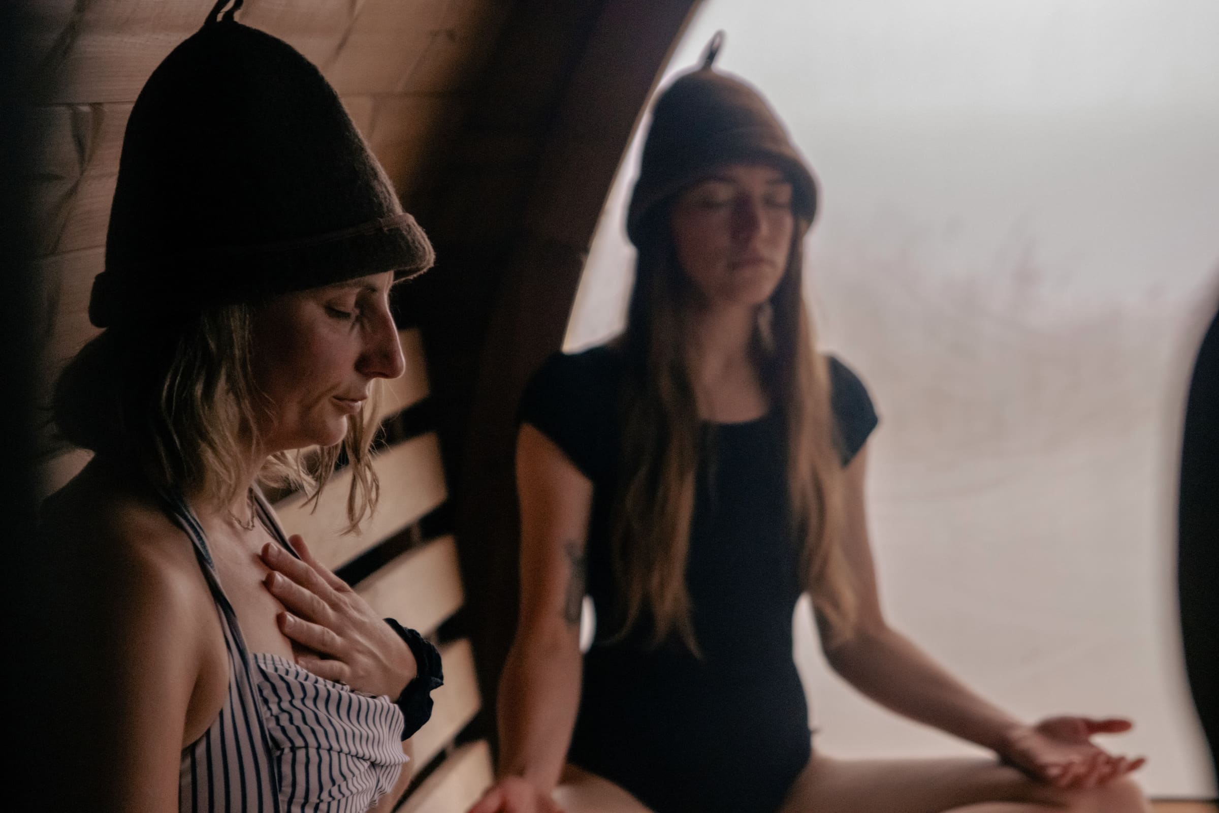 Two women meditating inside the Soul Sweats sauna, wearing felt sauna hats and sitting in calm, warm light.