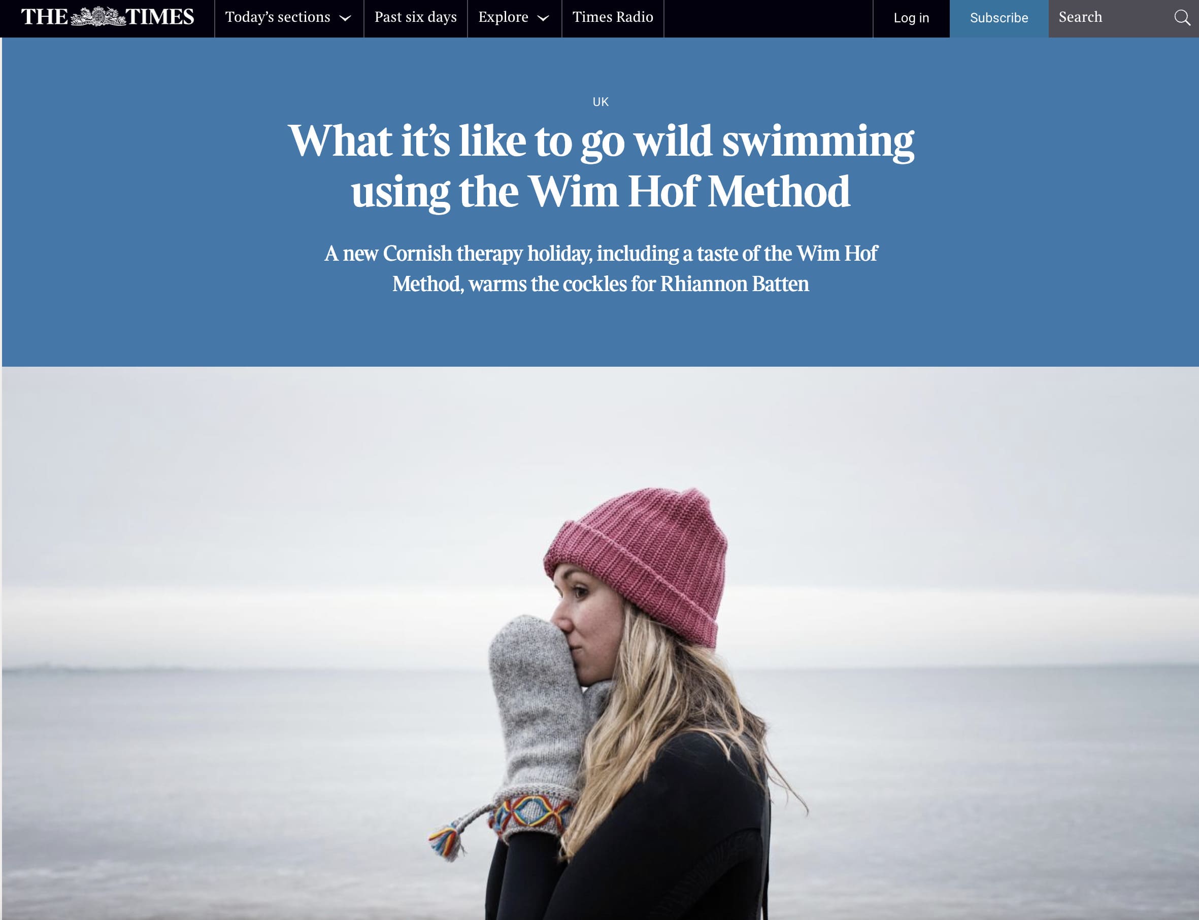 Woman standing on a calm shoreline wearing a pink beanie and grey mittens, featured in The Times article about wild swimming with the Wim Hof Method.