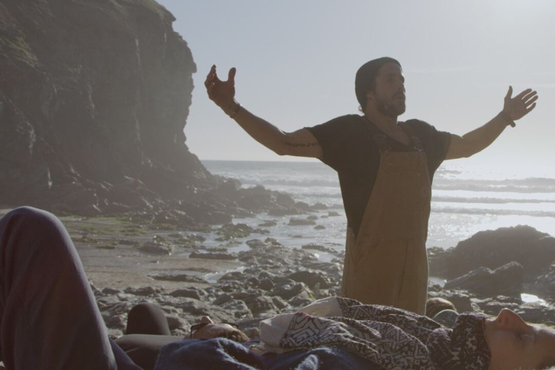 Man leading a breathwork session on a rocky beach at sunrise, with participants lying down in the foreground.