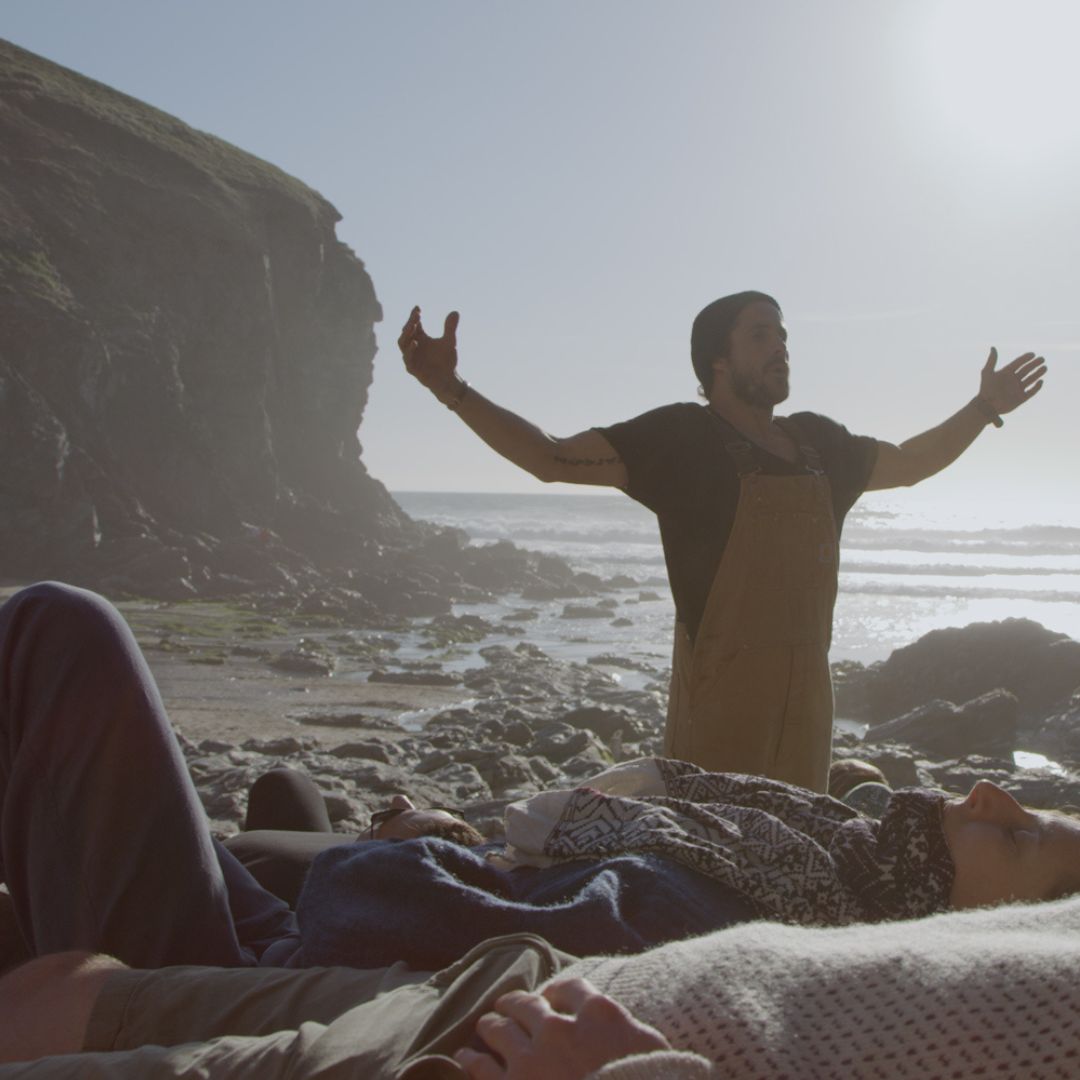 Man leading a breathwork session on a rocky beach at sunrise, with participants lying down in the foreground.